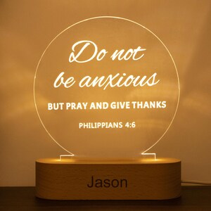 May include: A personalised night light featuring a wooden base and a circular acrylic top. The acrylic displays the text "Do not be anxious, but pray and give thanks, Philippians 4:6" and the name "Jason". The light emits a warm glow.