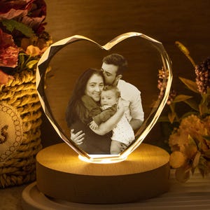 Puede incluir: Un marco de fotos de cristal en forma de corazón con un retrato familiar, iluminado por una luz cálida. El marco se apoya en una base de madera, rodeado de flores en una cesta y un jarrón. Un regalo único y personalizado.