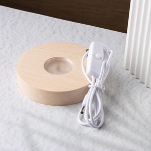 May include: A round, light-colored wooden LED light base with a clear, circular inset. A white power cord with an on/off switch is connected to the base. The base is on a light-colored surface.