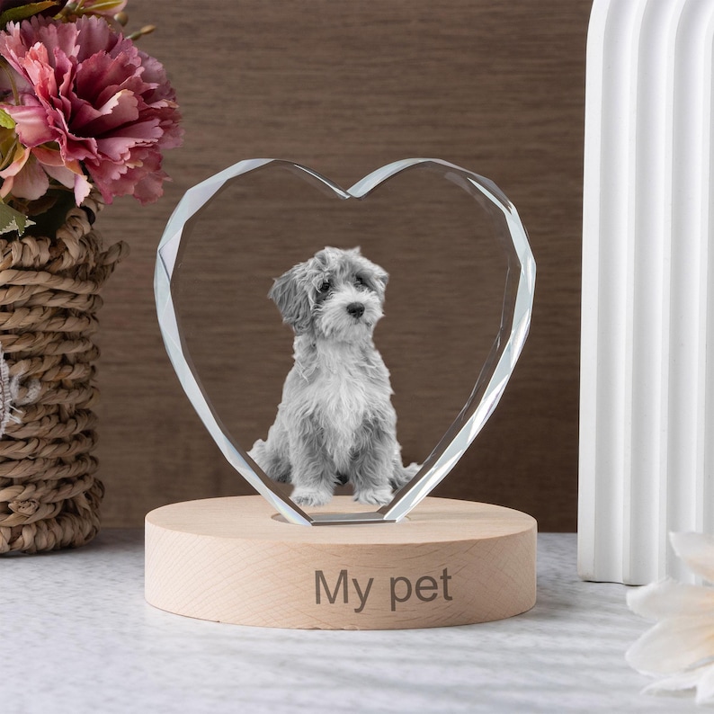 May include: A clear crystal heart-shaped ornament with a photo of a small, gray and white dog inside. The ornament sits on a round, light-colored wooden base with the words "My pet" engraved on it. The background includes flowers and a white decorative element.