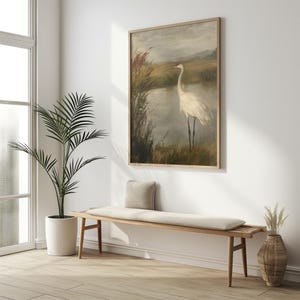 May include: A framed painting depicting a white crane standing in a marsh with reeds and water. The painting features a muted colour palette of browns, greens, and greys.