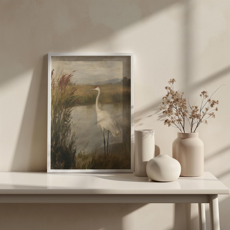May include: A framed print of a white bird standing in a marsh with reeds and a body of water. The print is in a white frame and is hanging on a white wall.