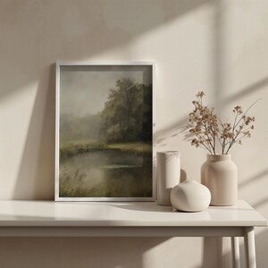 K&ouml;nnte beinhalten: Ein gerahmtes Landschaftsbild mit einem See, B&auml;umen und einer Holzbr&uuml;cke im Hintergrund. Das Bild h&auml;ngt an einer wei&szlig;en Wand &uuml;ber einem wei&szlig;en Tisch mit zwei beigefarbenen Keramikvasen und einer wei&szlig;en Keramik-Kugel.