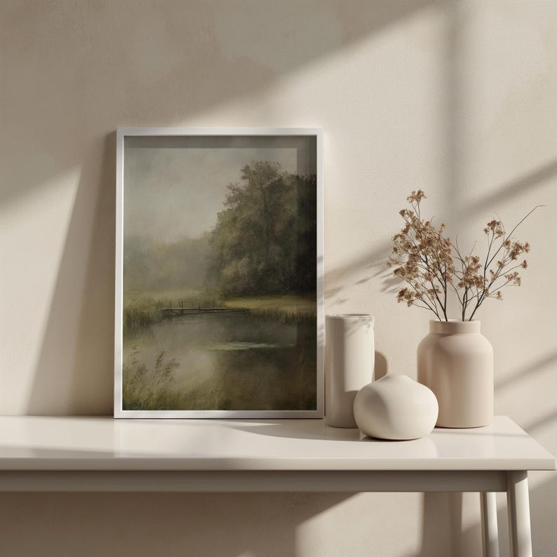 K&ouml;nnte beinhalten: Ein gerahmtes Landschaftsbild mit einem See, B&auml;umen und einer Holzbr&uuml;cke im Hintergrund. Das Bild h&auml;ngt an einer wei&szlig;en Wand &uuml;ber einem wei&szlig;en Tisch mit zwei beigefarbenen Keramikvasen und einer wei&szlig;en Keramik-Kugel.