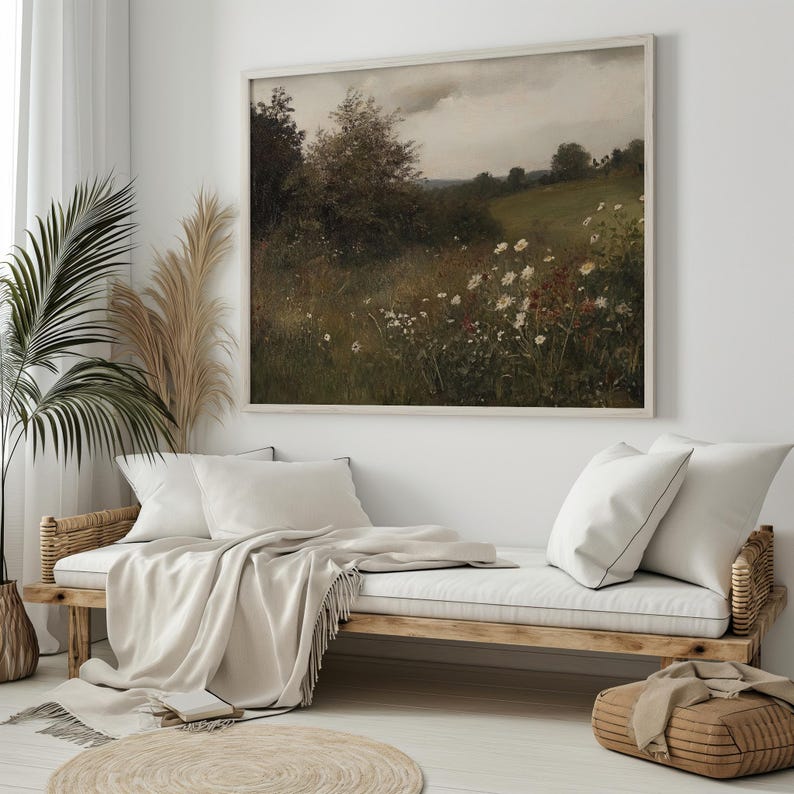 May include: A framed landscape painting depicts a field of wildflowers and trees under a cloudy sky. The artwork hangs above a wooden daybed with white pillows and a beige throw blanket. A woven basket and a round rug are in the foreground.