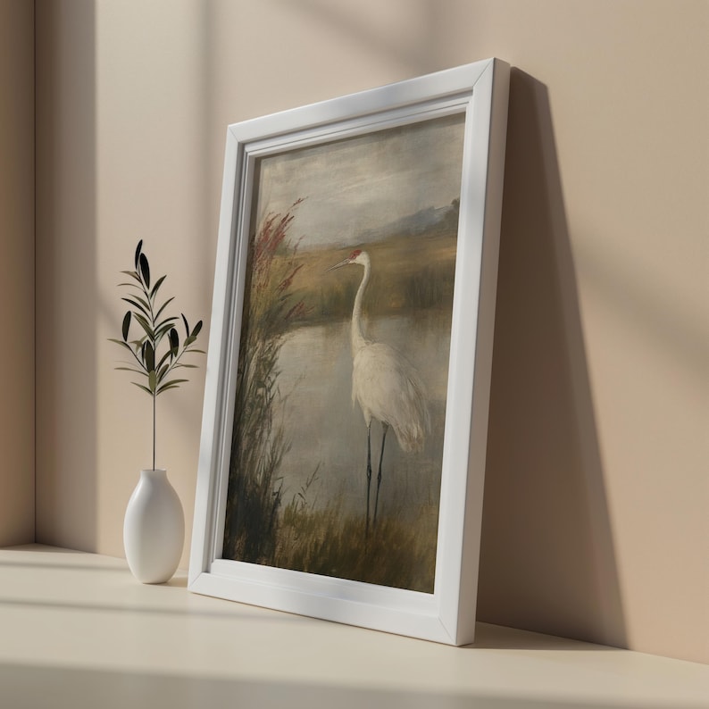 May include: A framed painting of a white crane standing in a marsh with reeds and water. The painting is in a white frame.