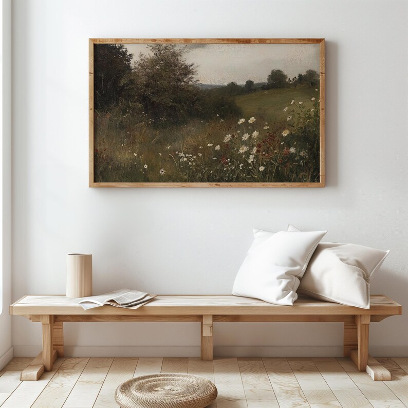 May include: A framed landscape painting depicts a field of wildflowers and trees under a cloudy sky. The artwork is in a wooden frame and hangs above a wooden bench with white pillows. A cylindrical vase and a rolled mat are also visible.