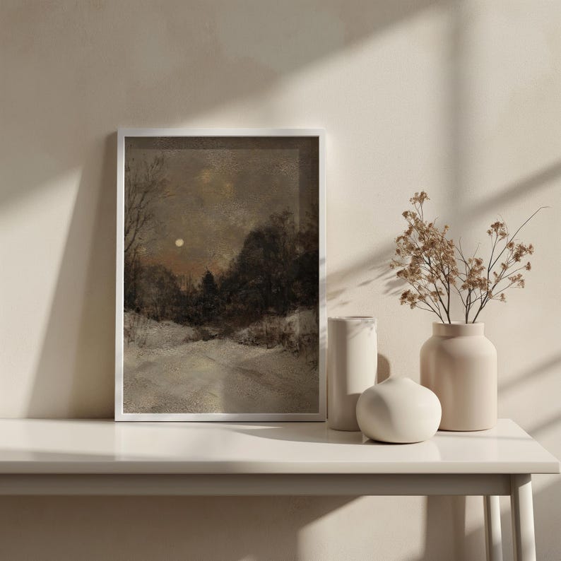 May include: A framed painting of a snowy landscape with a full moon in the sky. The painting is in a white frame and is sitting on a white table with two white vases and a white decorative object.