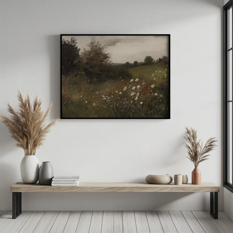 May include: A framed landscape painting depicts a field of wildflowers under a cloudy sky. The artwork features a meadow with white and red flowers, green grass, and trees. The painting is displayed above a wooden bench with decorative vases and books.
