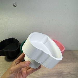 May include: A white, 3D-printed cookie cutter in the shape of a heart is held in a hand. Other cookie cutters in black, green, and pink are visible in the background. The cutter has a scalloped edge.