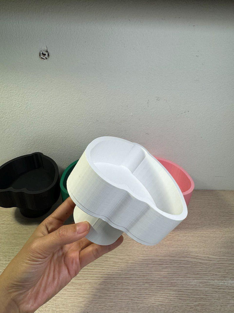May include: A white, 3D-printed cookie cutter in the shape of a heart is held in a hand. Other cookie cutters in black, green, and pink are visible in the background. The cutter has a scalloped edge.