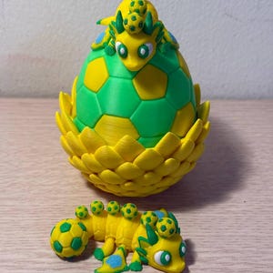May include: A yellow and green dragon sculpture with a soccer ball theme. The dragon is emerging from a soccer ball-shaped egg. A smaller dragon is lying down nearby. The sculpture is made of plastic.