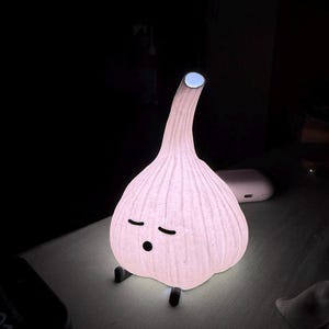 May include: A whimsical, light-up garlic bulb night light. The lamp is white with a textured surface and a small, smiling face. The stem has a blue light at the tip. The lamp is sitting on a white surface.