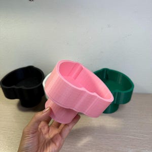 May include: A hand holds a pink heart-shaped container, with a black and green container in the background. The containers have a pedestal base and a heart-shaped bowl. The containers are made of plastic.