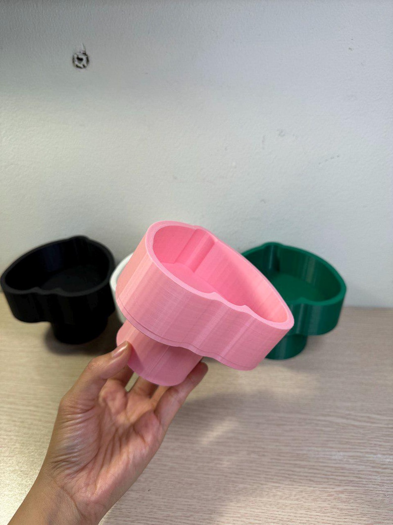 May include: A hand holds a pink heart-shaped container, with a black and green container in the background. The containers have a pedestal base and a heart-shaped bowl. The containers are made of plastic.