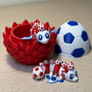 May include: A red, 3D-printed dragon-like creature with soccer ball accents, a red flower pot, and a soccer ball. The dragon has a white head, blue accents, and soccer balls on its back. The flower pot is red.