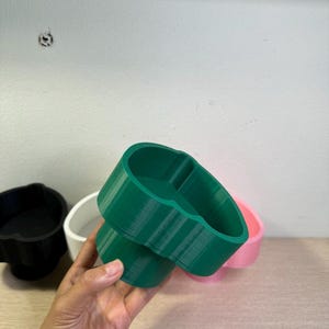 May include: A green, 3D-printed planter with a unique, rounded shape. The planter is held in a hand, with other planters in black, white, and pink visible in the background. The planter is made of plastic.