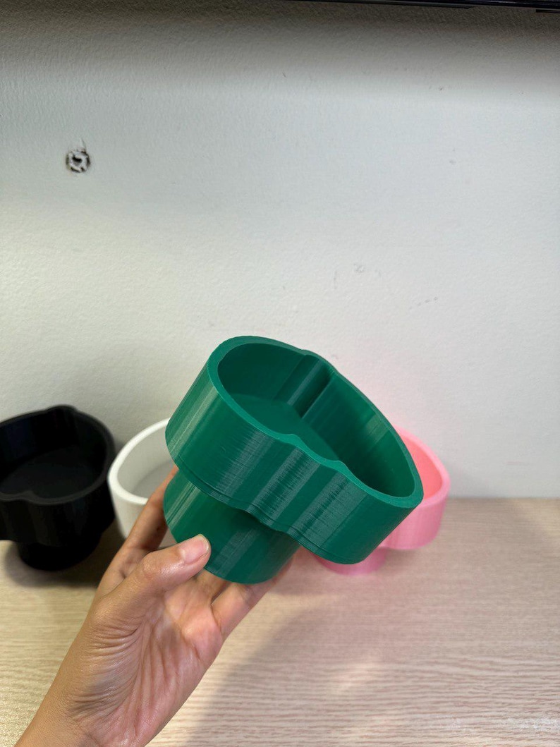 May include: A green, 3D-printed planter with a unique, rounded shape. The planter is held in a hand, with other planters in black, white, and pink visible in the background. The planter is made of plastic.