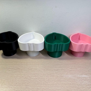 May include: Four heart-shaped containers in black, white, green, and pink. Each container has a scalloped edge and sits on a cylindrical base. The containers are arranged in a row on a light-colored wooden surface.