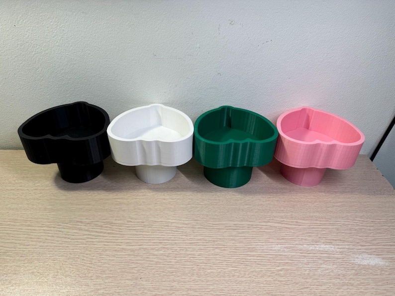 May include: Four heart-shaped containers in black, white, green, and pink. Each container has a scalloped edge and sits on a cylindrical base. The containers are arranged in a row on a light-colored wooden surface.