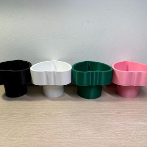 May include: Four heart-shaped cup holders in black, white, green, and pink. Each holder has a cylindrical base and a heart-shaped top, designed to hold cups or small items. The holders are arranged on a light brown wooden surface.