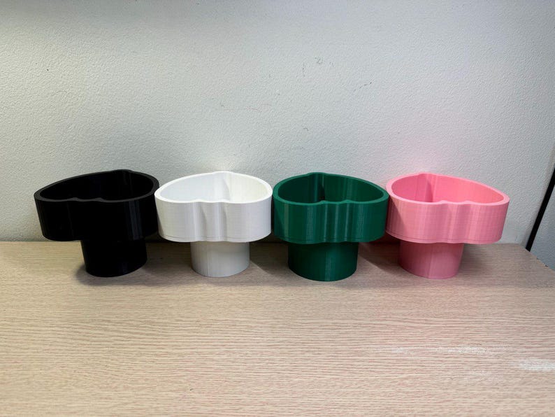 May include: Four heart-shaped cup holders in black, white, green, and pink. Each holder has a cylindrical base and a heart-shaped top, designed to hold cups or small items. The holders are arranged on a light brown wooden surface.