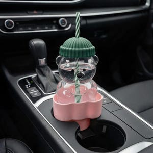 May include: A clear, bear-shaped water bottle with a green lid and a striped straw. The bottle is filled with water and sits in a pink cup holder. The cup holder is placed in a car's cup holder.