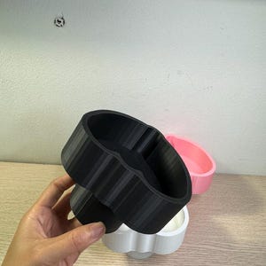 May include: A hand holds a black, 3D-printed, heart-shaped container with a ribbed exterior. A white and pink container are in the background. The containers are likely for holding small items or as decorative pieces.