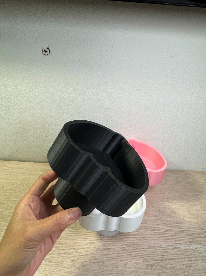 May include: A hand holds a black, 3D-printed, heart-shaped container with a ribbed exterior. A white and pink container are in the background. The containers are likely for holding small items or as decorative pieces.