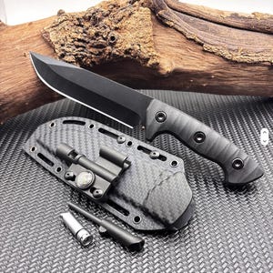 May include: A fixed-blade knife with a matte black finish and a textured black handle. The knife includes a carbon fibre patterned sheath equipped with a small integrated compass, a fire starter rod, and a separate metal striker tool.