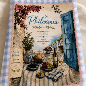 Könnte beinhalten: Ein hellblau-weiß kariertes Geschirrtuch mit einem Aufdruck einer mediterranen Szene, einer blauen Tür, einem Tisch mit Wein und Obst sowie dem Text Philoxenia, Kalis Eithate! You are welcome here! und verschiedenen Definitionen.