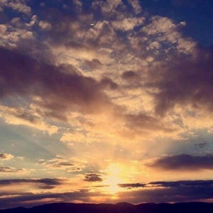 以下が含まれることがあります： 紫、ピンク、オレンジ、黄色の雲が混ざり合った、鮮やかな夕焼け空。太陽が雲の後ろに沈み、温かい光を放っています。