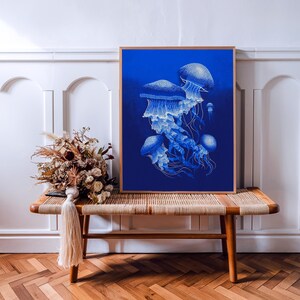 May include: A framed art print featuring a detailed illustration of several jellyfish in shades of blue and white against a deep blue background. The artwork is displayed on a wooden bench with a woven top, alongside dried flowers.