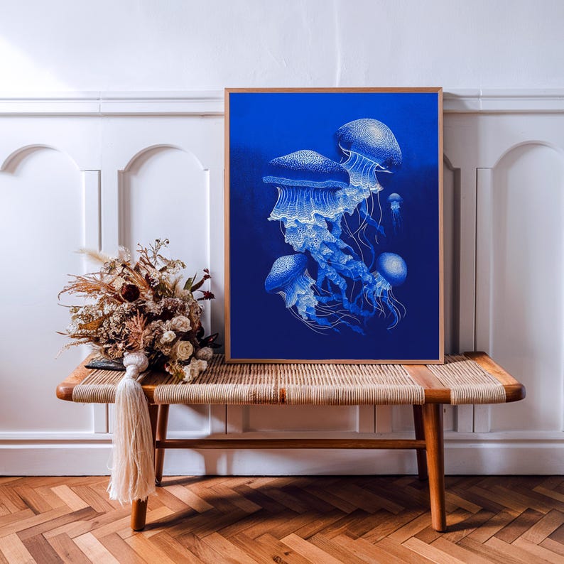 May include: A framed art print featuring a detailed illustration of several jellyfish in shades of blue and white against a deep blue background. The artwork is displayed on a wooden bench with a woven top, alongside dried flowers.