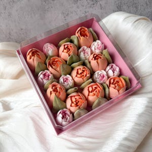 Pu&ograve; includere: Una scatola regalo quadrata rosa contenente un assortimento di dolcetti a forma di fiore nelle tonalit&agrave; pesca, arancione e rosa chiaro, accentuati da foglie verdi. La scatola &egrave; coperta da un coperchio in plastica trasparente e strutturata.