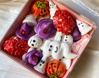 Può includere: Una scatola rosa piena di dolcetti a tema Halloween. I dolcetti includono meringhe bianche a forma di fantasma, meringhe di zucca arancioni, meringhe a forma di cappello da strega viola e meringhe rosse a forma di cervello. La scatola è su una superficie bianca.