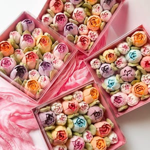 Pu&ograve; includere: Quattro scatole quadrate in plastica trasparente riempite con un assortimento di dolciumi multicolori a forma di fiore. I dolcetti sono disposti in grappoli compatti, con sfumature di rosa, viola, arancione, giallo e blu, accentuati da foglie verdi.