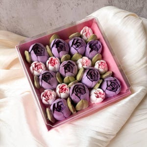 Pu&ograve; includere: Una scatola quadrata in plastica trasparente contenente un assortimento di dolciumi a forma di fiore nelle tonalit&agrave; del viola scuro, lavanda chiaro e rosa tenue, intervallati da piccole foglie verdi, disposti in un vassoio interno rosa.