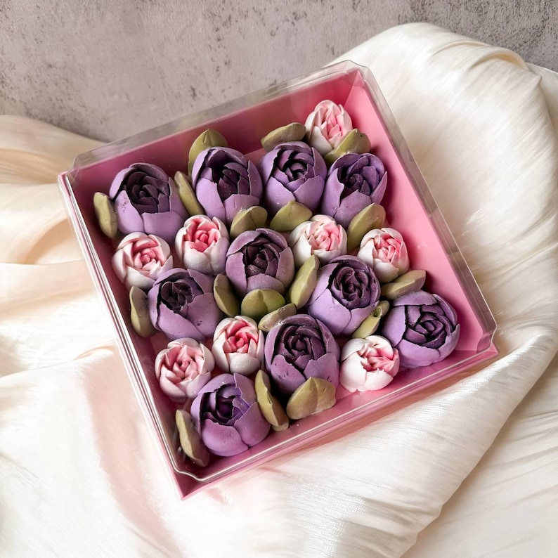 Pu&ograve; includere: Una scatola quadrata in plastica trasparente contenente un assortimento di dolciumi a forma di fiore nelle tonalit&agrave; del viola scuro, lavanda chiaro e rosa tenue, intervallati da piccole foglie verdi, disposti in un vassoio interno rosa.