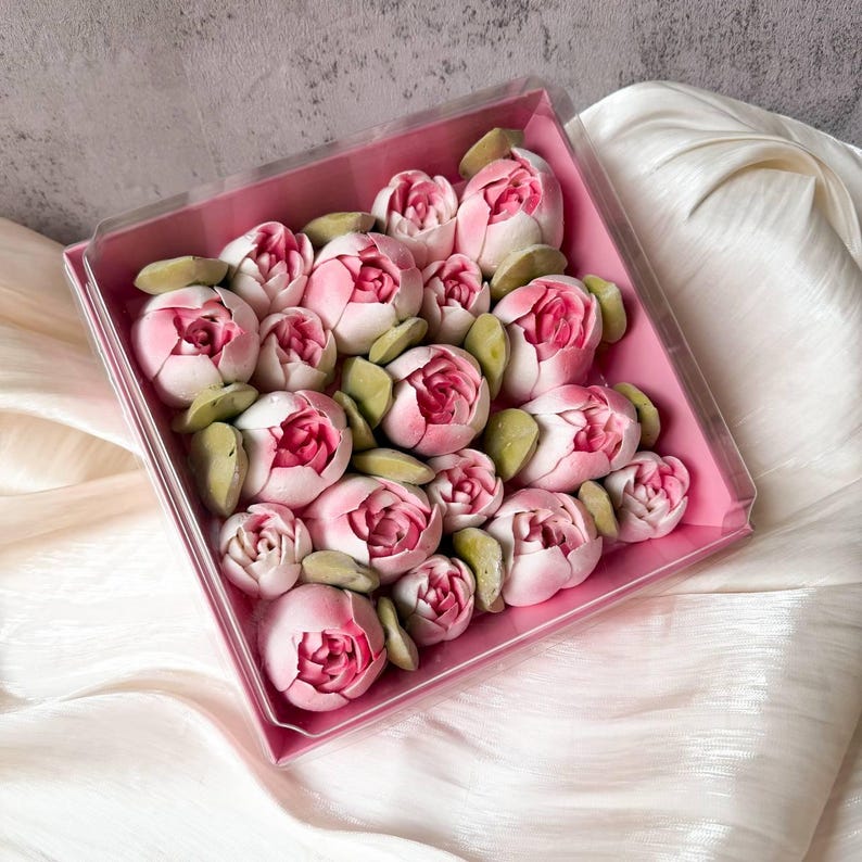 Pu&ograve; includere: Una scatola quadrata rosa contenente numerosi piccoli dolciumi arrotondati a forma di fiore, nei toni del rosa chiaro e scuro, intervallati da piccoli pezzi a forma di foglia di colore verde pallido. La scatola ha un coperchio in plastica trasparente.