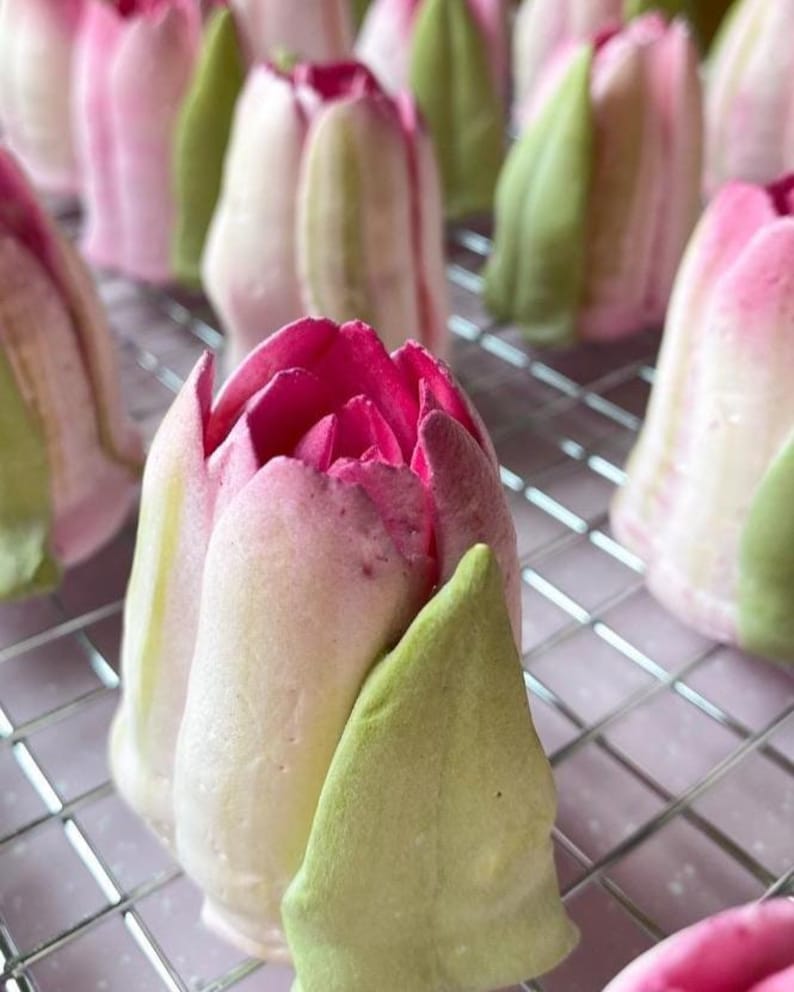 Pu&ograve; includere: Una collezione di dolciumi a forma di tulipano disposti su una griglia metallica. Ogni pezzo presenta una base rosa pallido e bianca con un centro magenta intenso e una singola foglia verde chiaro attaccata al lato, imitando una flor.