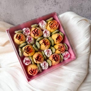 Pu&ograve; includere: Una scatola regalo quadrata rosa contenente un assortimento di dolciumi a forma di fiore. La composizione presenta diversi fiori simili a rose arancioni e gialle intervallati da boccioli rosa pallido e foglie verde chiaro, il tutto sotto un coperchio trasparente.
