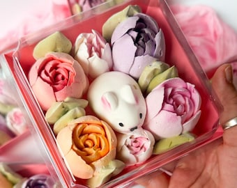 May include: A clear plastic box containing a collection of molded marshmallow confections shaped like various flowers in shades of pink, orange, purple, and white, alongside a central white marshmallow rabbit with pink-tipped ears and black eyes.