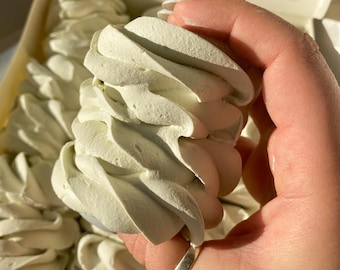 May include: Close-up of a light green, swirled dessert held in a hand. The dessert has a soft, textured appearance and is part of a larger collection in a white box. The dessert is a light green color.