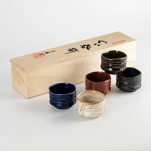 May include: A set of six square ceramic sake cups in various colors, including blue, brown, black, and white. The cups have a textured, organic design. They are presented with a wooden box that has Japanese text and a red stamp.