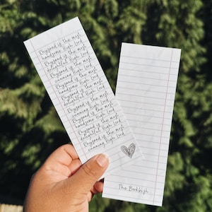 May include: Two white paper bookmarks with lined paper design. One bookmark has the text "Rhysand is the most handsome High Lord" repeated multiple times. The other bookmark is blank. A small heart is drawn on one bookmark, and "The Bookbish" is written on the other.
