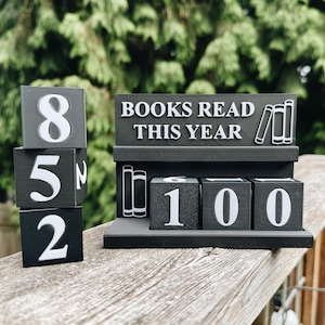 May include: A black wooden book counter with white numbers and the text "BOOKS READ THIS YEAR." The counter displays "852" on stacked blocks and "100" on the base. Decorative bookshelf icons are also present.