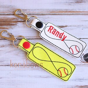 May include: Two personalized keychains with a baseball design. One keychain is white with the name "Randy" embroidered in red and a red baseball. The other keychain is yellow with a black infinity symbol and a red baseball.