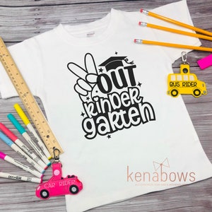 May include: White t-shirt with black text that says "Out Kinder Garten" with a peace sign and graduation cap. The shirt is on a wooden surface with a pink car keychain and a yellow school bus keychain.