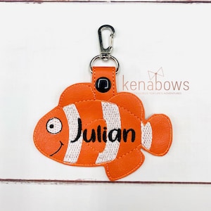 May include: Orange and white clownfish keychain with the name Julian embroidered in black. The keychain has a silver clip and a black button.